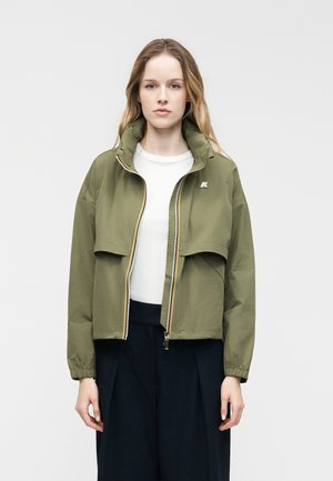 Νεαρή γυναίκα που κοιτάζει μπροστά, φορώντας ένα ανοικτό χακί μπουφάν πάνω από ένα λευκό τοπ και σκούρα φαρδιά παντελόνια, μπροστά σε απλό φόντο.