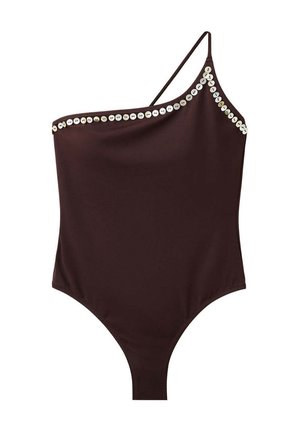 Mujer con el pelo mojado, vistiendo un bañador negro de un solo hombro con adornos circulares, de pie frente a un fondo rocoso a la luz del sol.