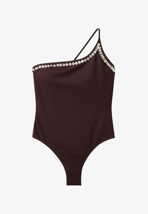 Mujer con el pelo mojado, vistiendo un bañador negro de un solo hombro con adornos circulares, de pie frente a un fondo rocoso a la luz del sol.