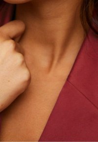 Gros plan sur un décolleté en forme de V profond, mettant en valeur une peau lisse et un tissu bordeaux doux en superposition. La main repose légèrement sur la clavicule.