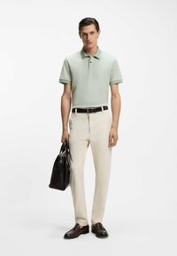 Hellgrünes Polohemd mit kurzen Ärmeln, cremefarbene Hose, dunkelbrauner Gürtel und passende Loafers. Trägt eine schwarze Ledertasche.