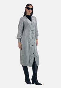 Virkat grå kofta med ett kabelmönster, oversized ärmar, framknappar och en djup slits; kombinerat med svarta åtsittande byxor och spetsiga stövlar.