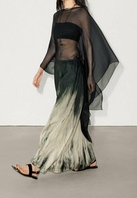 Cape transparent noir sur un crop top noir ajusté, associé à une jupe ombragée verte fluide. Des sandales plates noires complètent le look.