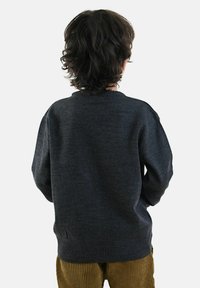 Bambino con capelli ricci scuri, indossa un maglione grigio scuro e pantaloni in velluto a coste marroni, rivolto di spalle su uno sfondo chiaro e semplice.