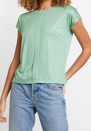 Kobieta w miętowo-zielonym topie z krótkim rękawem i detalem szwu z przodu oraz jasnoniebieskich, wysokim stanem dżinsach, z ręką spoczywającą w kieszeni.