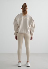 Beige oversize mikina s vyšitým nápisem "AIM'N APPAREL" na zadní straně, spojená s obtáhnutými béžovými legínami a bílými sportovními botami.