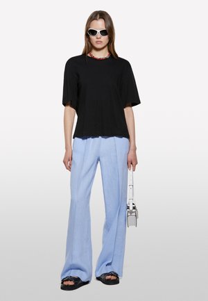 T-shirt oversized noir, pantalon large bleu clair, lunettes de soleil blanches, sandales noires avec des accents, et une ceinture blanche avec un petit sac.