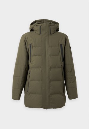Homme portant une doudoune vert olive les mains dans les poches, pantalon noir et chaussures noires, debout devant un fond blanc.