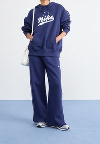 Tmavě modrá mikina s kapucí a bílým logem Nike, doplněná o široké kalhoty stejné barvy a vícebarevné sportovní boty. Viditelný detail bílé kabelky.