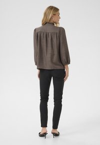 Blouse longue marron texturée à col volanté et dos froncé, associée à un pantalon noir slim et des mules noires à talons.