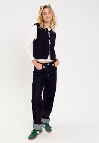 Zwarte mouwloze vest met een strik aan de voorkant, gedragen over een witte blouse met lange mouwen. Gecombineerd met donkere jeans en groene sneakers met een gumsole.