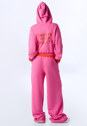 Persona con felpa con cappuccio e pantaloni rosa, con la scritta "CREATE YOUR REALITY" sulla felpa, vista di spalle su sfondo bianco.