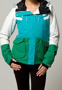 Giacca multicolore con cappuccio nero, corpo turchese e verde, due grandi tasche frontali e tasche pettorali con zip, realizzata in un materiale morbido.