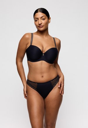 Vrouw die een zwart gewatteerd beha met dubbele bandjes en bijpassende slip met kanten rand draagt, poserend tegen een effen lichte achtergrond.