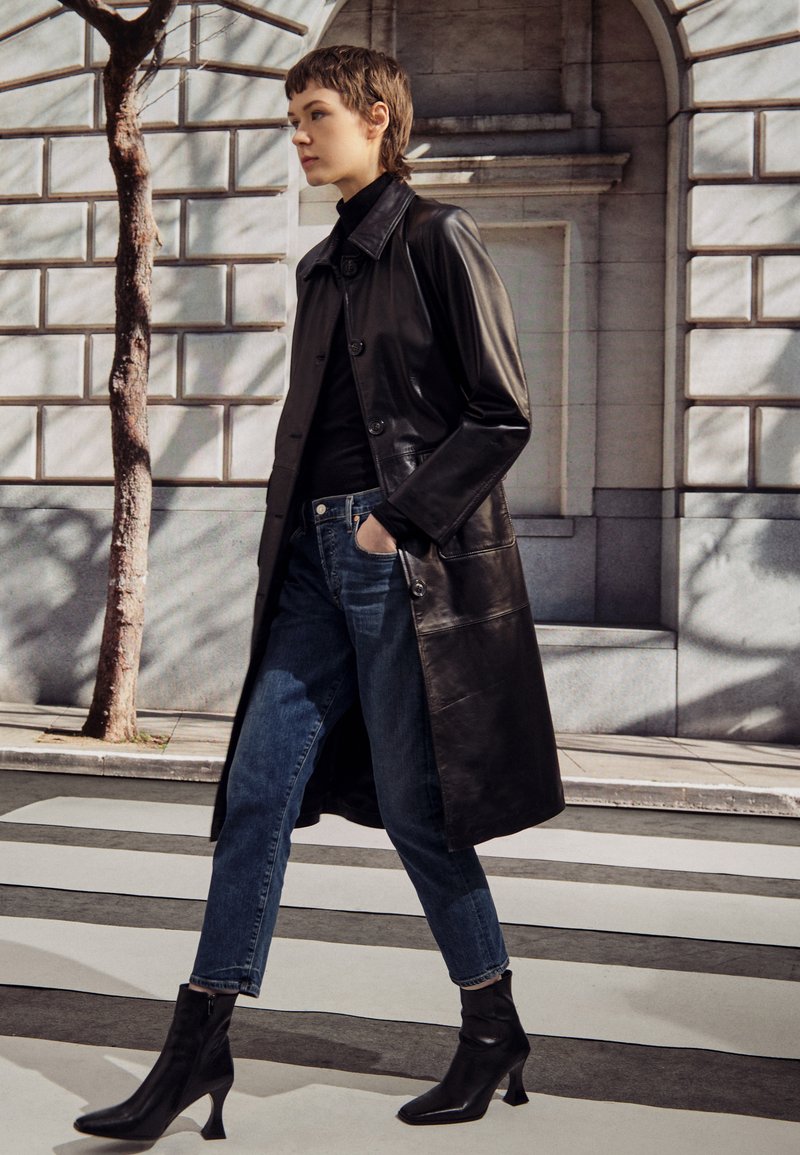 Schwarzer Leder-Trenchcoat mit Knöpfen, über einem schwarzen Rollkragenpullover gestylt. Kombiniert mit blauen Jeans und schwarzen Ankle-Boots auf einem Zebrastreifen.