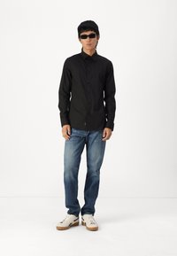 Camisa negra de manga larga con botones, jeans azules y zapatillas blancas con suelas marrones; el portador también lleva gafas de sol negras.