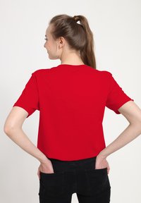 Młoda kobieta z długim brązowym kucykiem, ubrana w czerwoną koszulkę i czarne dżinsy, z rękami w tylnej kieszeni, odwrócona tyłem na białym tle.