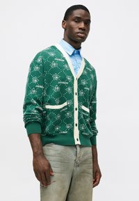 Cardigã verde com um acabamento em creme, apresentando padrões de personagens de cartoon e a palavra "SORVETE". Inclui dois bolsos frontais.