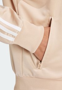 Béžová mikina s texturovanou tkaninou, obsahující kapsu na zip a bílé pruhované detaily na rukávech.