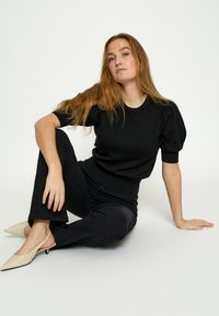 Jonge vrouw met lang rood haar zit op de vloer, draagt een zwarte top met korte mouwen en opgezette mouwen, zwarte broek en beige puntige schoenen.