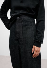 Korkeavyötäröiset mustat farkut, joissa on teksturoitu pinta, hienovaraiset ompeleet, sivutaskut ja napitus. Yhdistetty mustan neuleen kanssa.