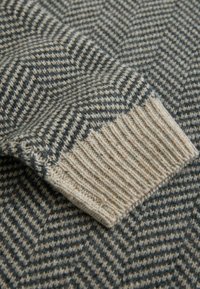 Houndstooth-patterned sweater in gray and beige wool with a ribbed beige cuff. The texture is soft and warm, suitable for cooler weather.