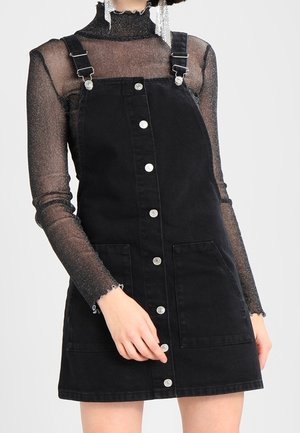 Mujer vestida con un vestido tipo pichi de denim negro con botones en la parte delantera sobre una blusa negra transparente de manga larga y brillante con pendientes plateados colgantes.