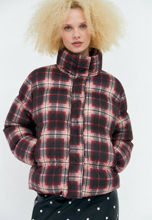 Donna con capelli biondi ricci che indossa una giacca imbottita a quadri rossa e nera e pantaloni neri a pois, in piedi con le mani in tasca.