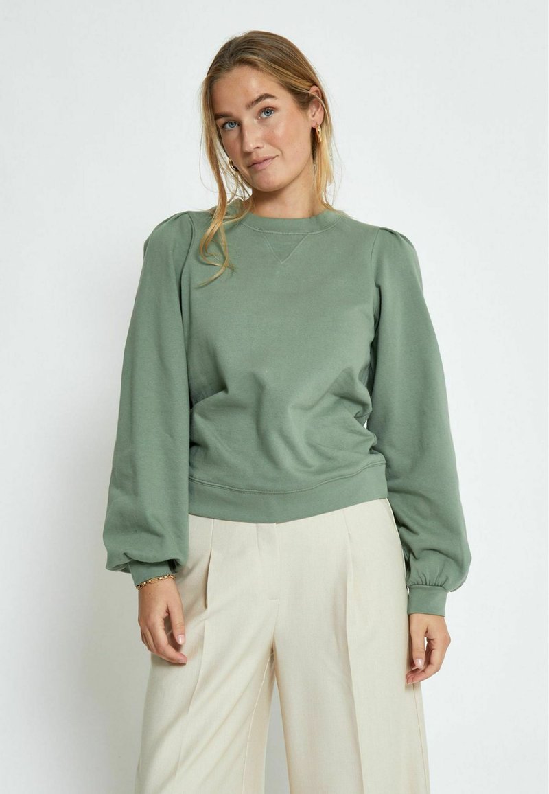 Green pullover sweatshirt with puffed sleeves, ribbed cuffs, and hem. Soft texture, crew neckline, and relaxed fit. Paired with light-colored trousers.