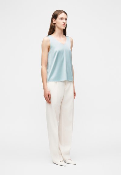 Νεαρή γυναίκα στέκεται φορώντας αμάνικη ανοιχτό μπλε μπλούζα, λευκά φαρδιά παντελόνια και λευκά παπούτσια με μυτερή μύτη, μπροστά σε ένα απλό φόντο.