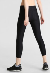 Leggings neri con una texture liscia, vita alta e lunghezza cropped, abbinati a sneakers nere con un accento colorato.