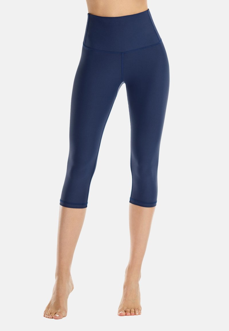 Frau trägt marineblaue, hoch taillierte, verkürzte Leggings und steht barfuß vor einem schlichten hellen Hintergrund.