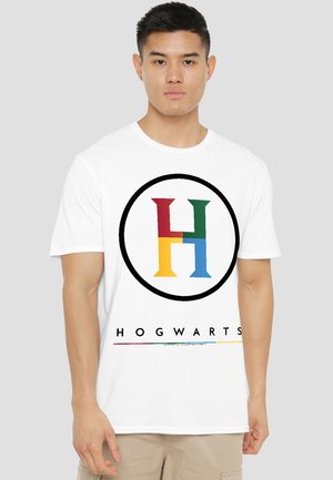 Young man wearing white T-shirt with multicolored "H" logo in circle and "Hogwarts" text below on plain background.