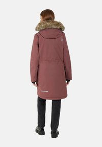 Parka lunga bordeaux con collo in pelliccia sintetica, materiale impermeabile, tasche laterali e dettaglio riflettente all'orlo. Pantaloni neri e scarpe robuste.