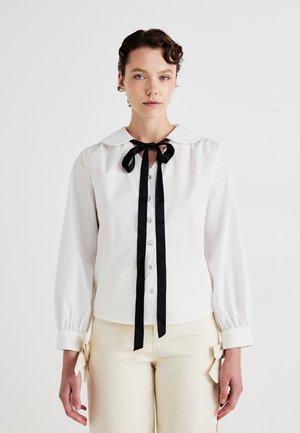 Mujer con una blusa blanca de botones y gran cuello, corbata negra tipo lazo, mangas largas y pantalones crema con lazos laterales.