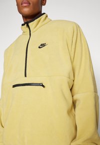 Gul fleecetröja med hög krage, svart dragkedja och ficka med dragkedja fram. Nike-logotyp i svart på bröstet.
