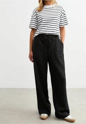 Person wearing a white and black striped T-shirt tucked into loose black drawstring pants with beige and white sneakers.