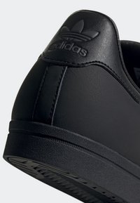 Talon de basket Adidas noir avec logo tréflé embossé et semelle texturée sur un fond clair.