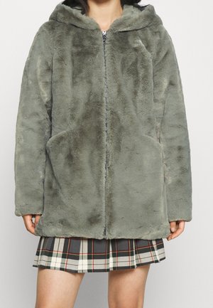 Personne portant un manteau à capuche en fausse fourrure gris-vert avec une fermeture éclair à l'avant et des poches, associé à une jupe plissée à carreaux noir, blanc et rouge.