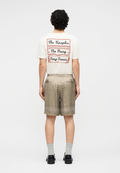 Homme aux cheveux bouclés portant un t-shirt blanc avec un texte rouge et un short beige à motifs, debout de dos sur un fond blanc.