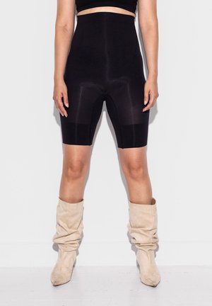 Kvinde iført højtaljede sorte cykelshorts og beige slaskede knælange støvler, stående foran en hvid baggrund.