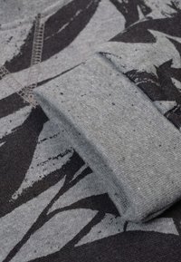 Grijze sweatshirtstof met zwart abstract patroon, met een getextureerde manchet en zichtbare stiksel details. Zacht, rekbaar materiaal zichtbaar.
