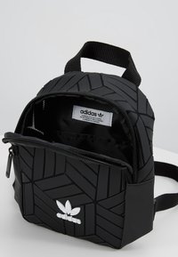 Svart Adidas mini-ryggsäck med geometriskt mönster, dragkedja, vitt logo, texturerat material och justerbar rem.