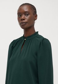 Long-sleeve dark green blouse, features ruffled neckline, keyhole opening, soft texture, and subtle gold button detail.