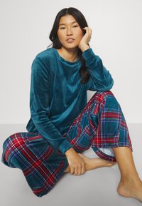 Top en velours teal à manches longues associé à un pantalon large à carreaux rouges et bleus, mettant en avant des textures douces et des motifs contrastés.