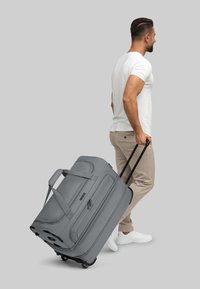 Grau gefärbte Reisetasche mit zwei ausziehbaren Griffen, robustem Stoff und Seitentaschen; neben einer Person in beigen Hosen und weißen Sneakers platziert.