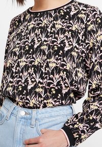 Blusa floreale nera con un motivo floreale rosa e giallo, maniche lunghe con polsini elasticizzati, abbinata a jeans a vita alta di colore blu chiaro.