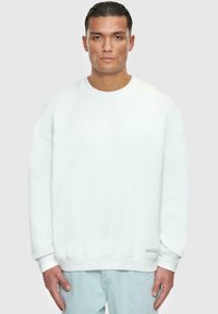 Sweatshirt blanc avec un col rond, des manches longues et des poignets côtelés. Présente une petite étiquette au bas de l'ourlet. Texture douce.