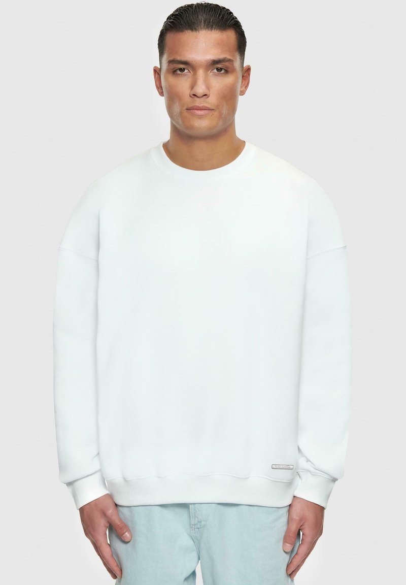 Sweatshirt blanc avec un col rond, des manches longues et des poignets côtelés. Présente une petite étiquette au bas de l'ourlet. Texture douce.