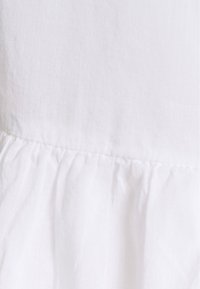 Valkoinen puuvillakangas, jossa on teksturoitu, hieman rypistynyt pinta. Alaosassa on rypytetty yksityiskohta.